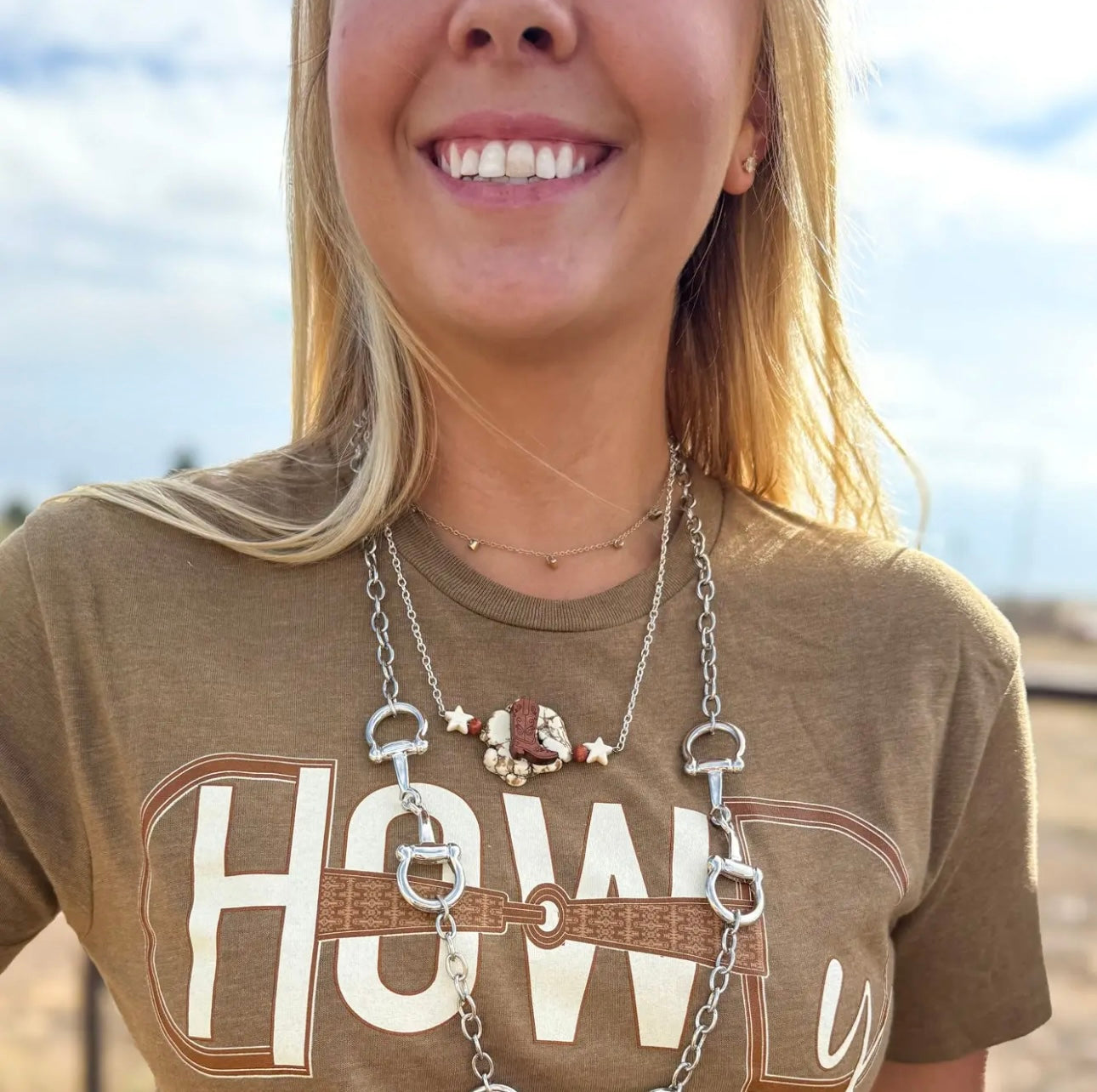 Person wearing a necklace with a horse bit charm, smiling outdoors.