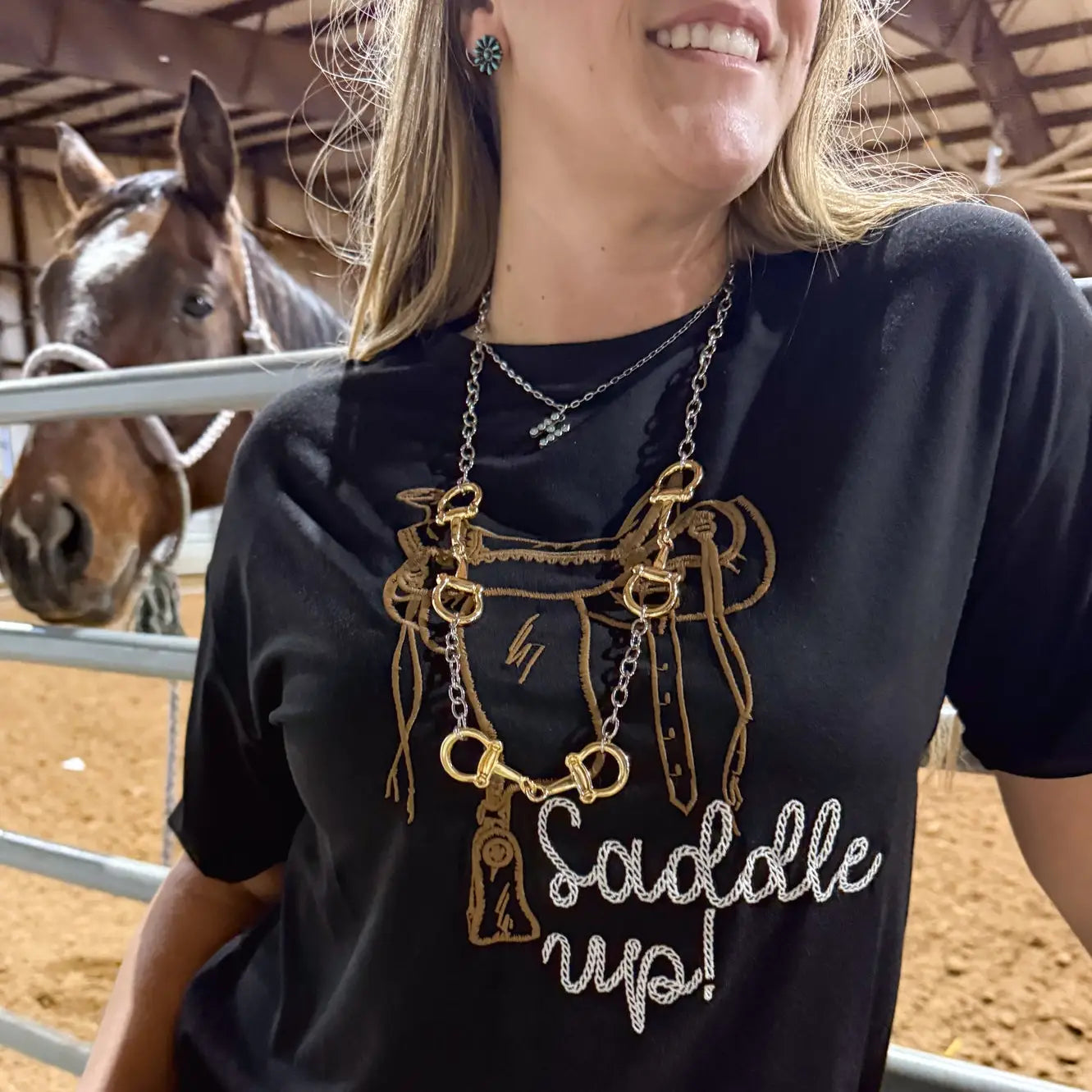 Person wearing a black t-shirt with a horse design and 'Saddle Up!' text, standing next to a horse in a stable.