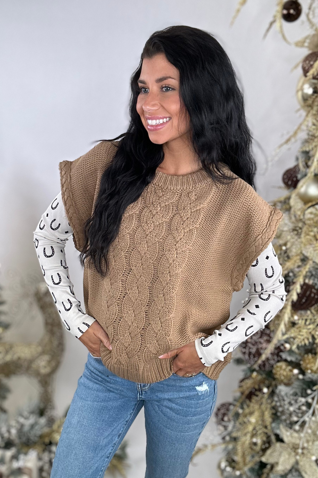 Woman wearing a brown cable knit sweater vest over a horseshoe patterned shirt with a neutral background.