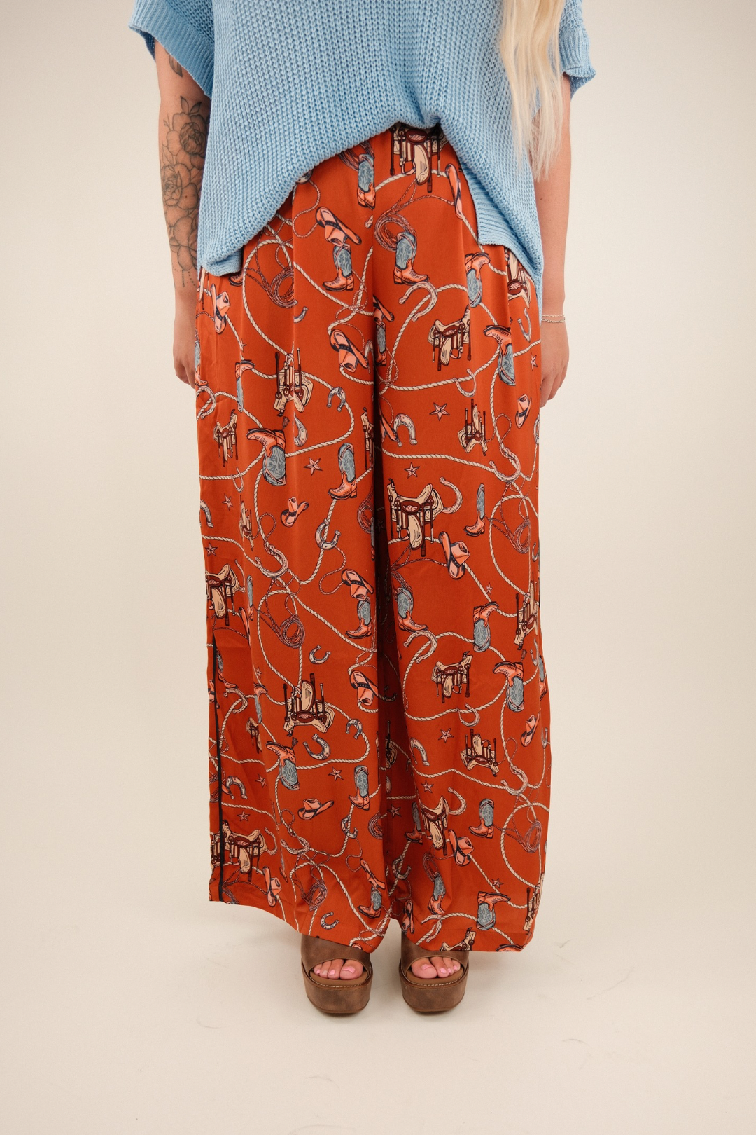 Rust orange wide-leg trousers with a high-waisted fit, made from silky faux silk fabric. Features an all-over vintage western print of cowboy boots, saddles, ropes, and stars.