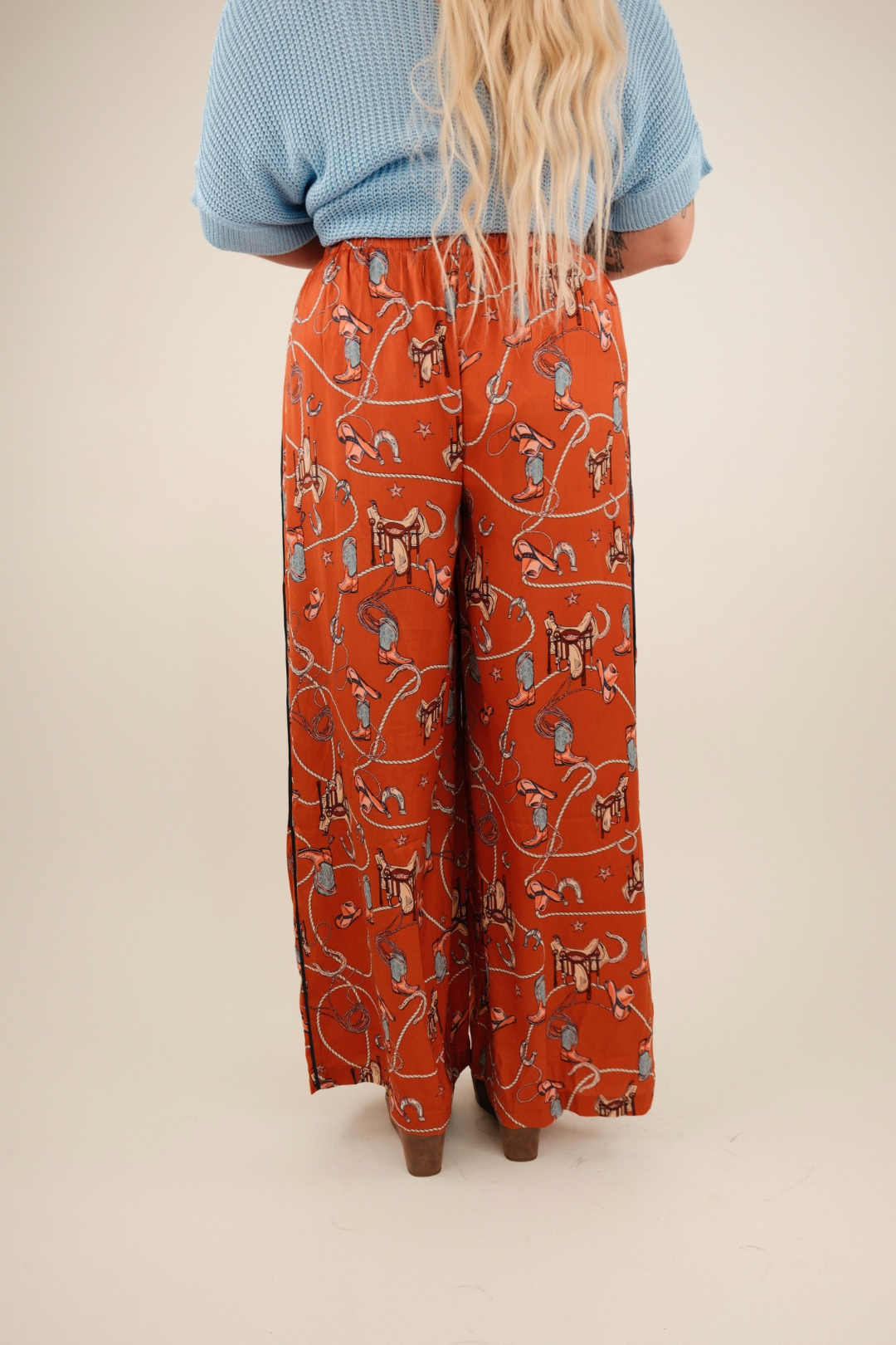 Rust orange silky wide-leg trousers with a high waist and vintage western print of boots, saddles, ropes, and stars.