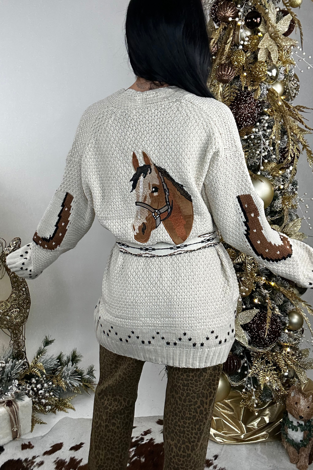 Person wearing a beige belted cardigan with a horse design across the back and horseshoes on the elbows.