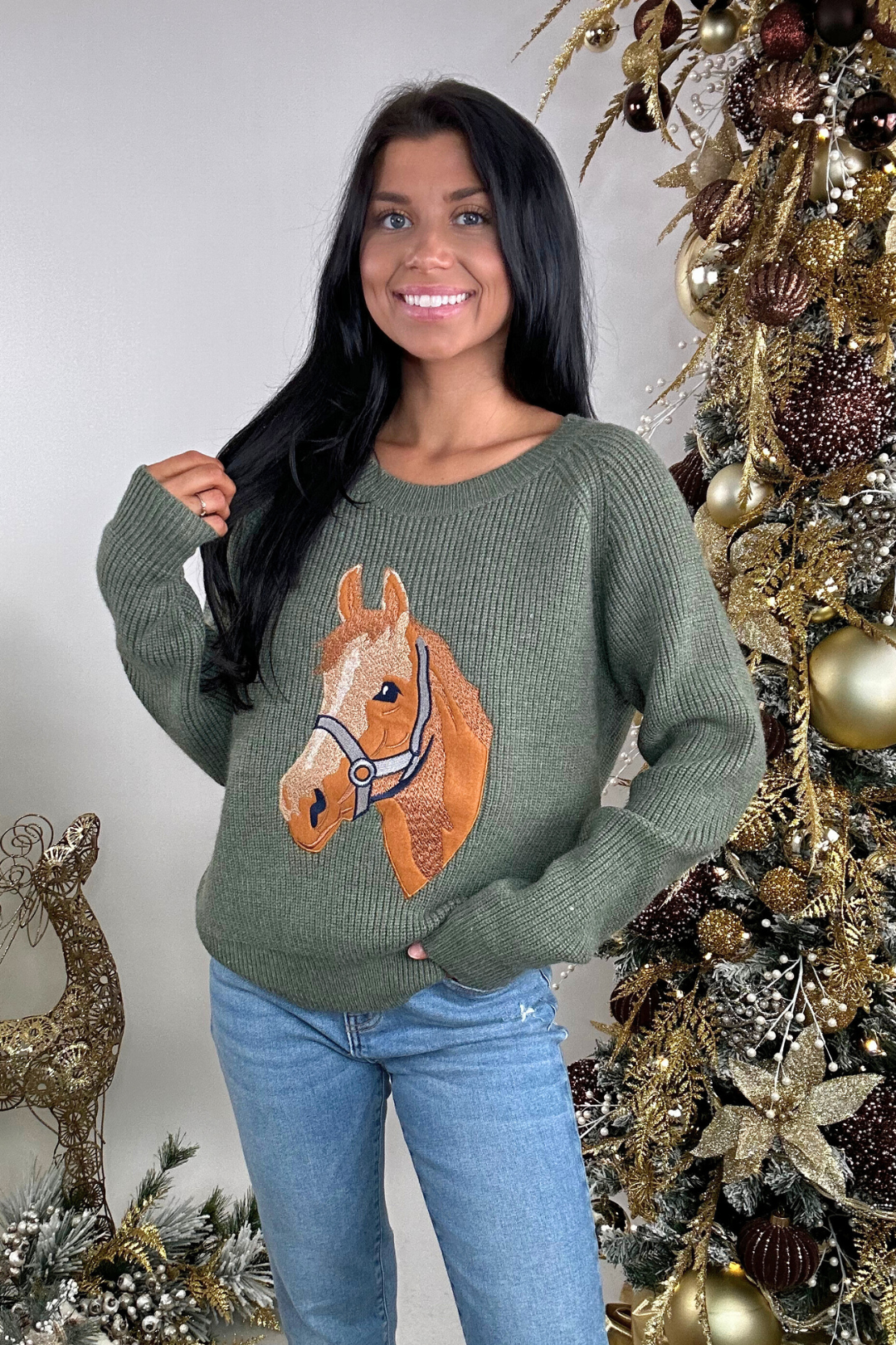 Woman wearing a green sweater with a horse design in front of a decorated Christmas tree.