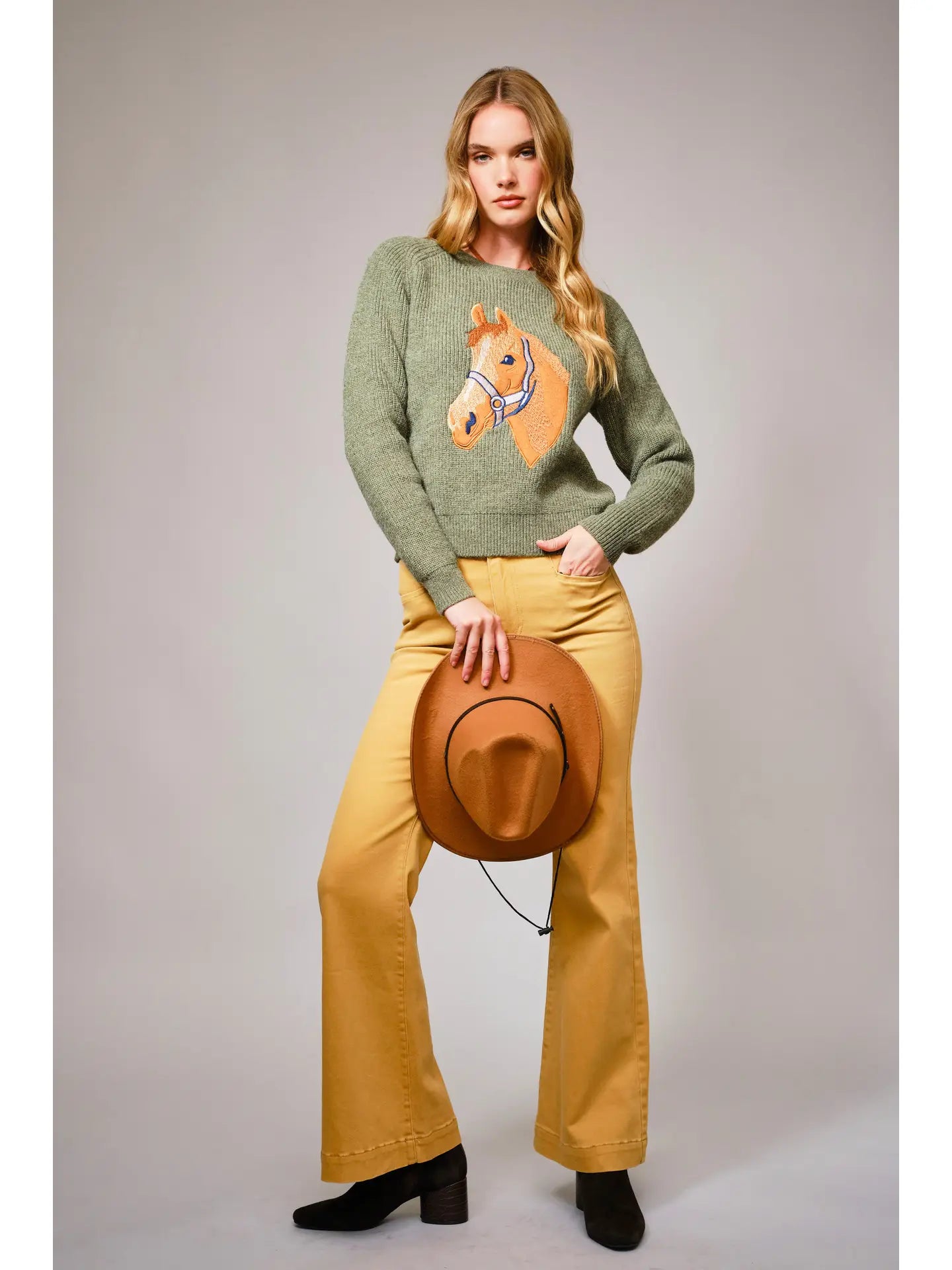 Woman wearing a green sweater with a horse design, yellow pants, and holding a brown hat against a gray background