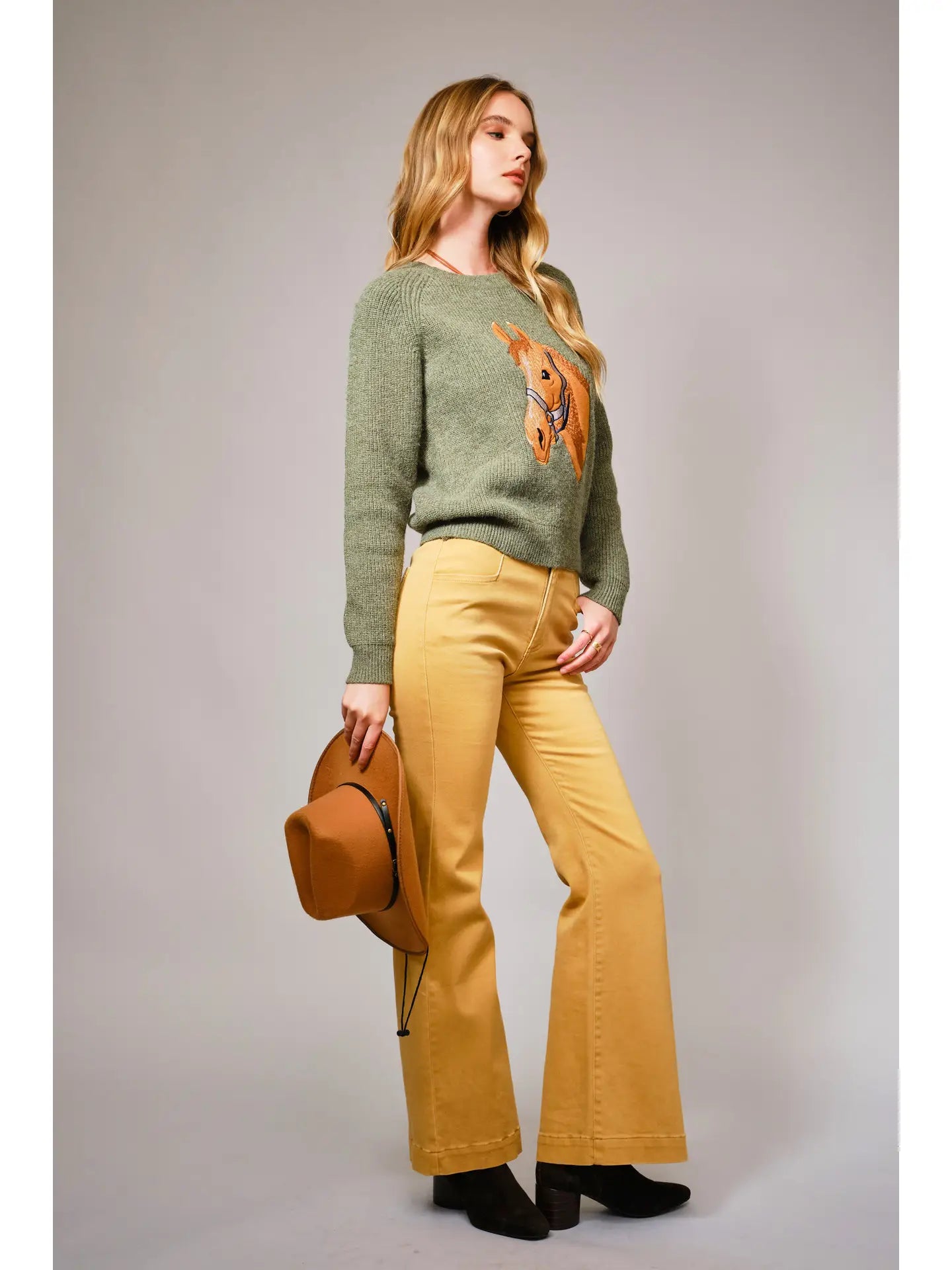Woman wearing a green sweater with a graphic horse design and yellow pants, holding a brown hat against a gray background.