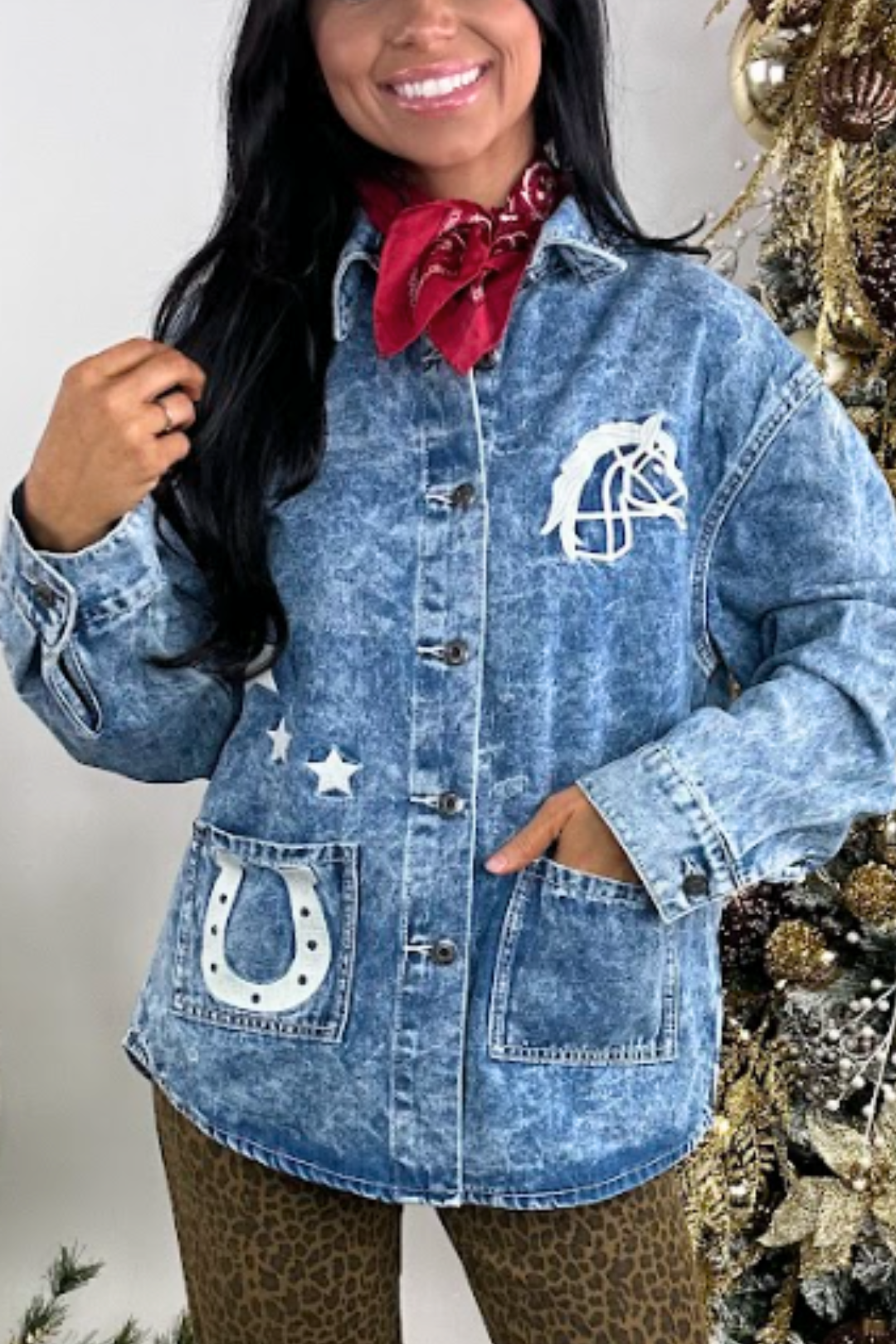 Person wearing a blue denim jacket with horse and star designs, standing next to a decorated Christmas tree.
