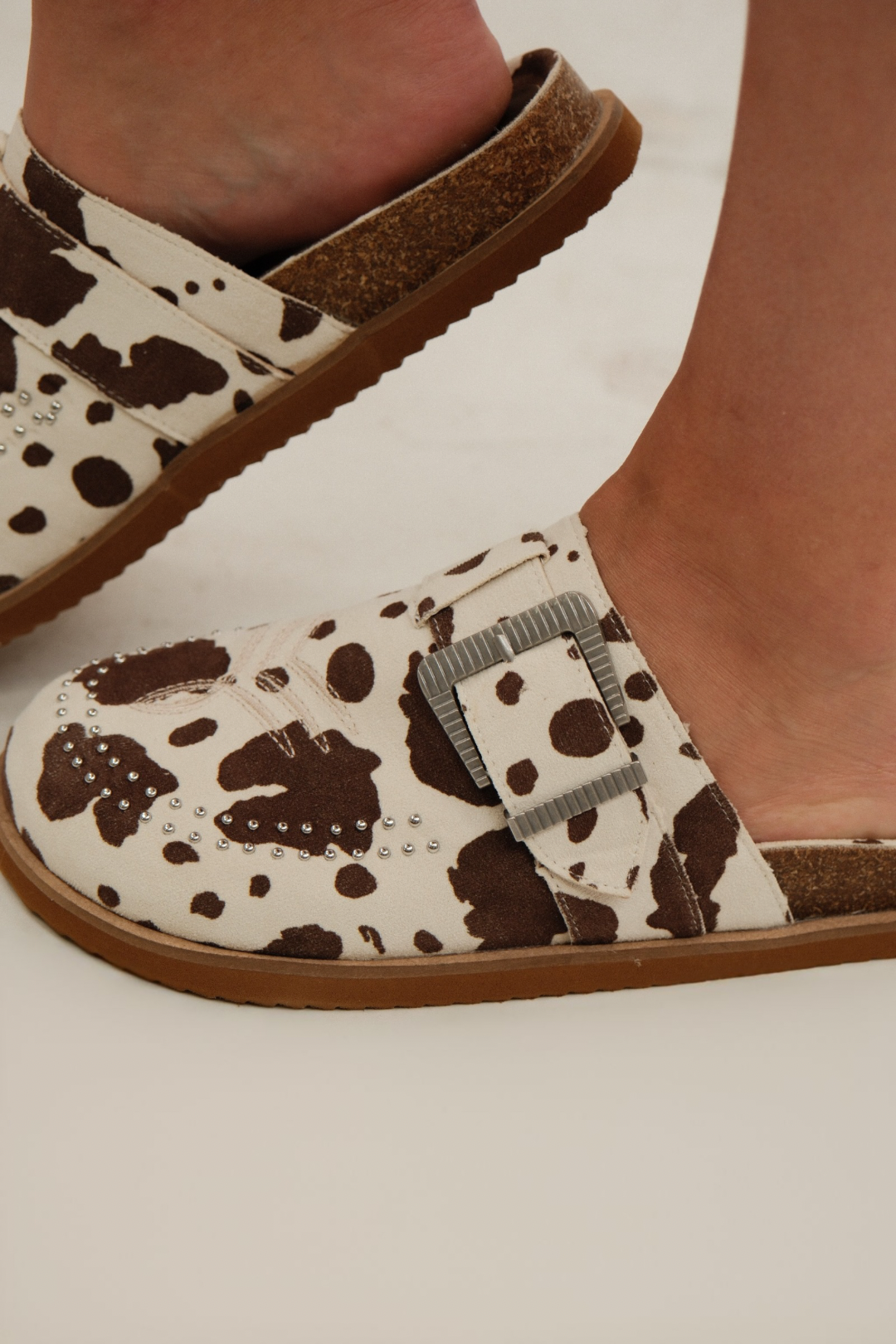 Side profile of Studded Rodeo Slides showing cow-print suede, western toe stitching and studded trim.