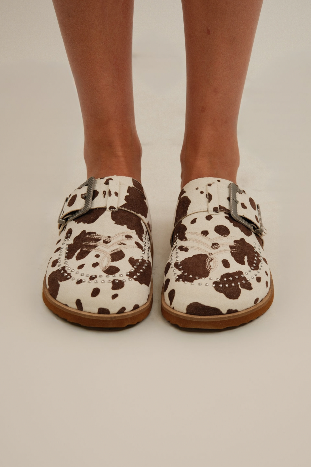 Front view of Studded Rodeo Slides, white and brown cow-print suede with western embroidery and silver studs.