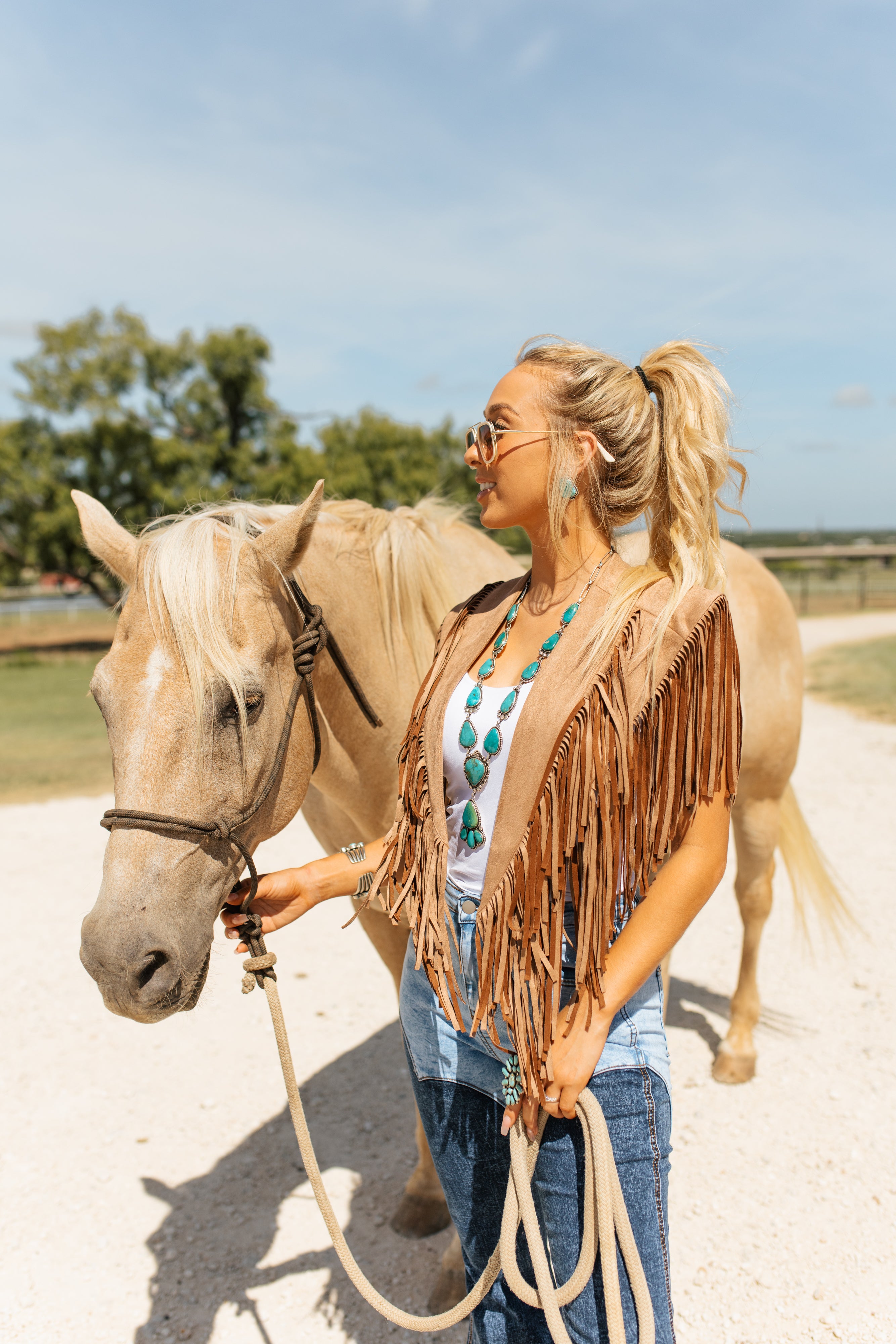 Woman in a fringe vest standing next to a horse on a dirt path with trees in the background