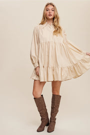 Woman wearing a beige dress with long sleeves and ruffled hem, paired with brown knee-high boots on a beige background.