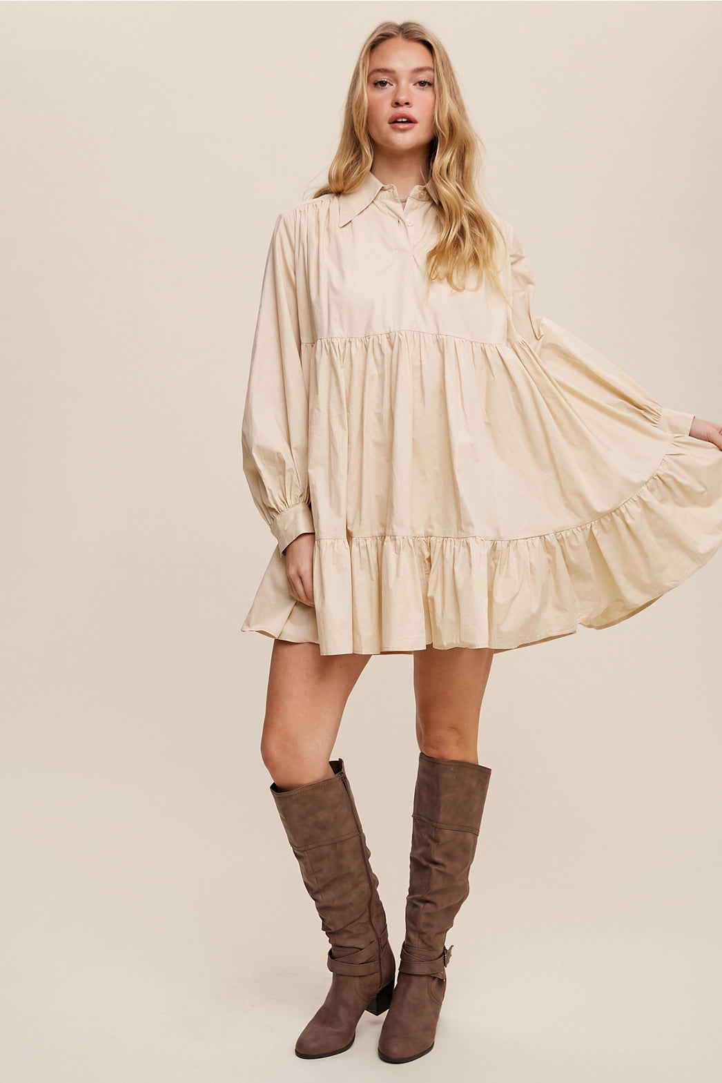 Woman wearing a beige dress with long sleeves and ruffled hem, paired with brown knee-high boots on a beige background.