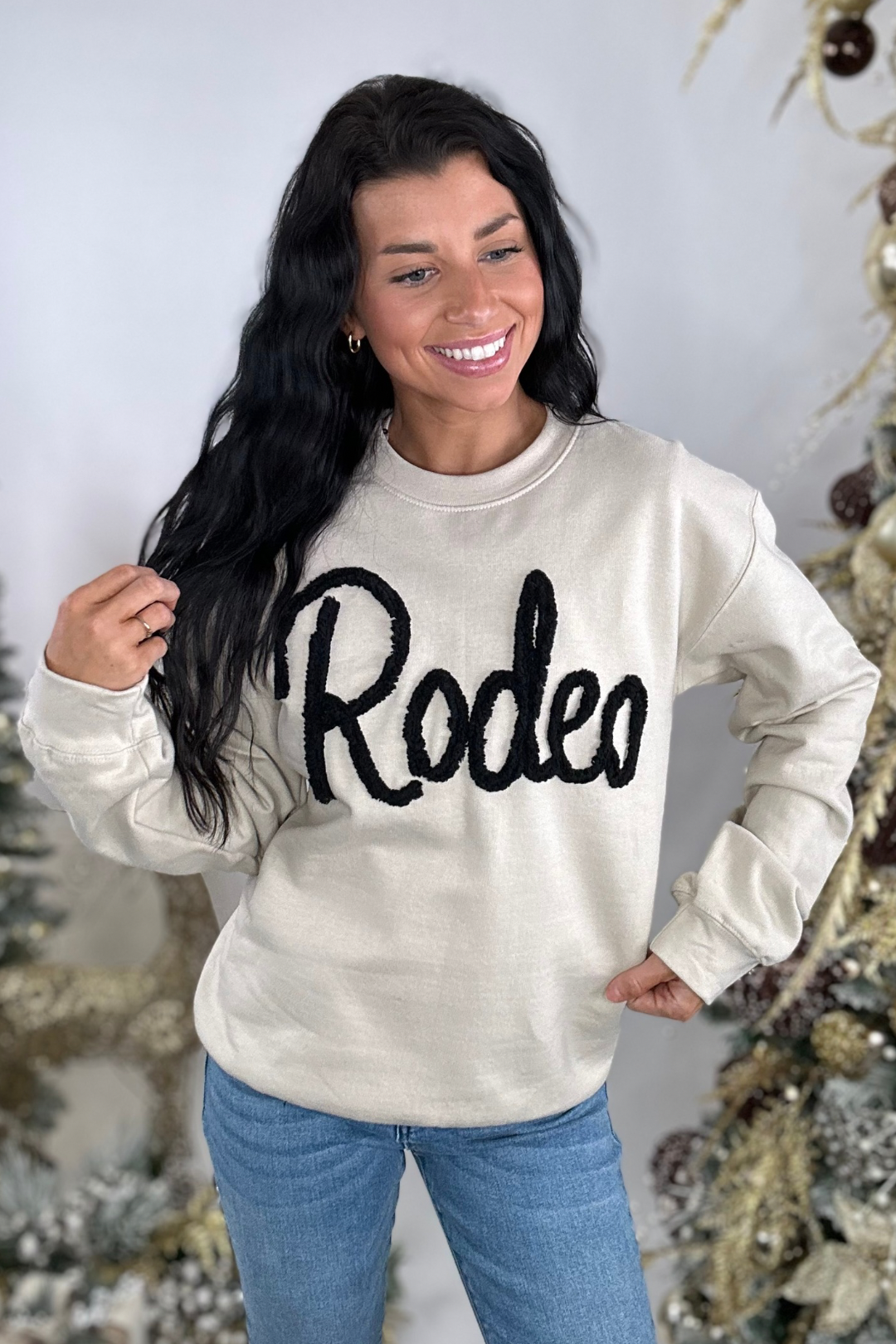 Woman wearing a beige sweatshirt with 'Rodeo' text, standing in front of a decorated Christmas tree.
