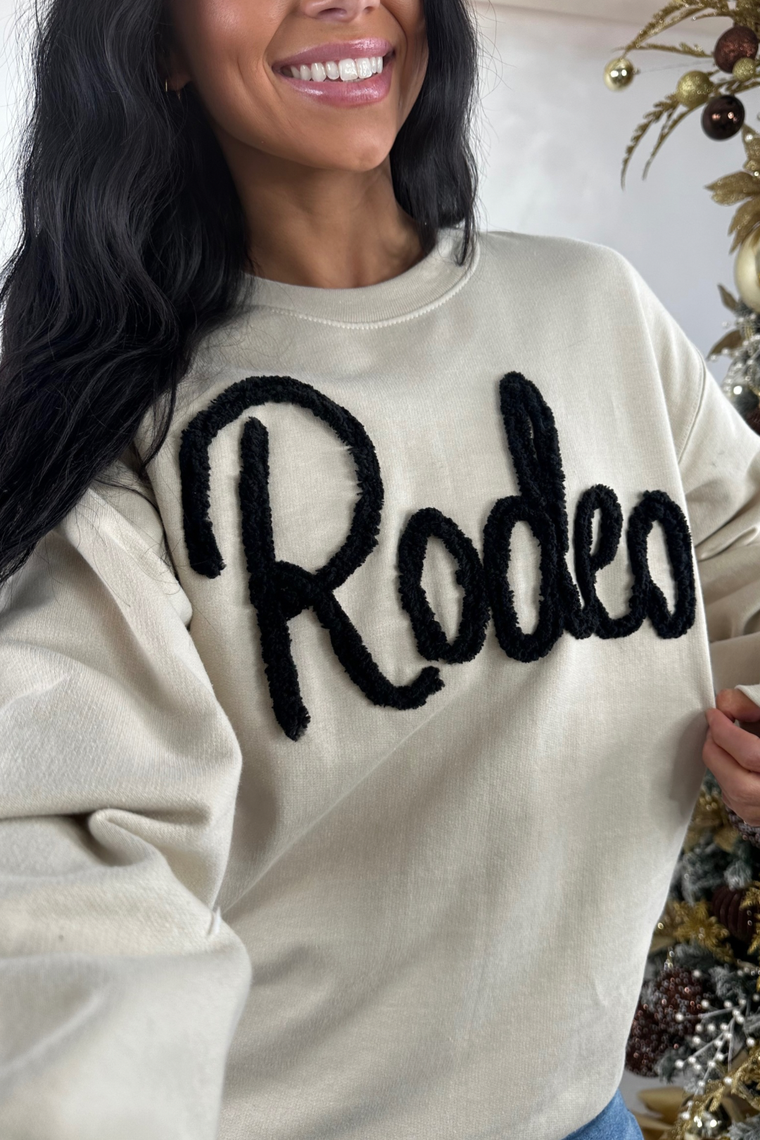 Person wearing a beige sweatshirt with 'Rodeo' text in black, standing in front of a decorated Christmas tree.