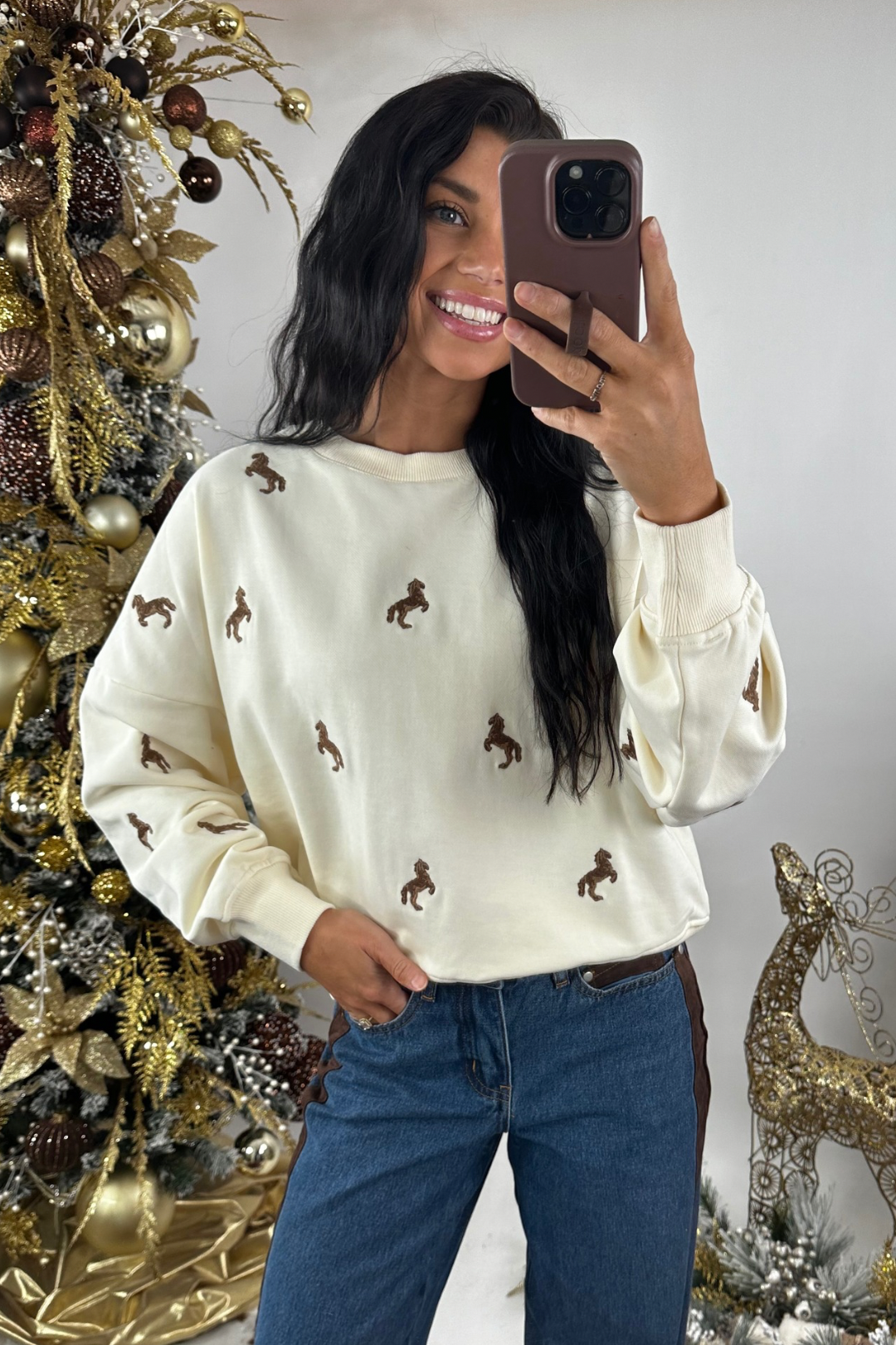 Woman taking a mirror selfie wearing a cream sweater with horse prints and blue jeans, standing next to a decorated Christmas tree.
