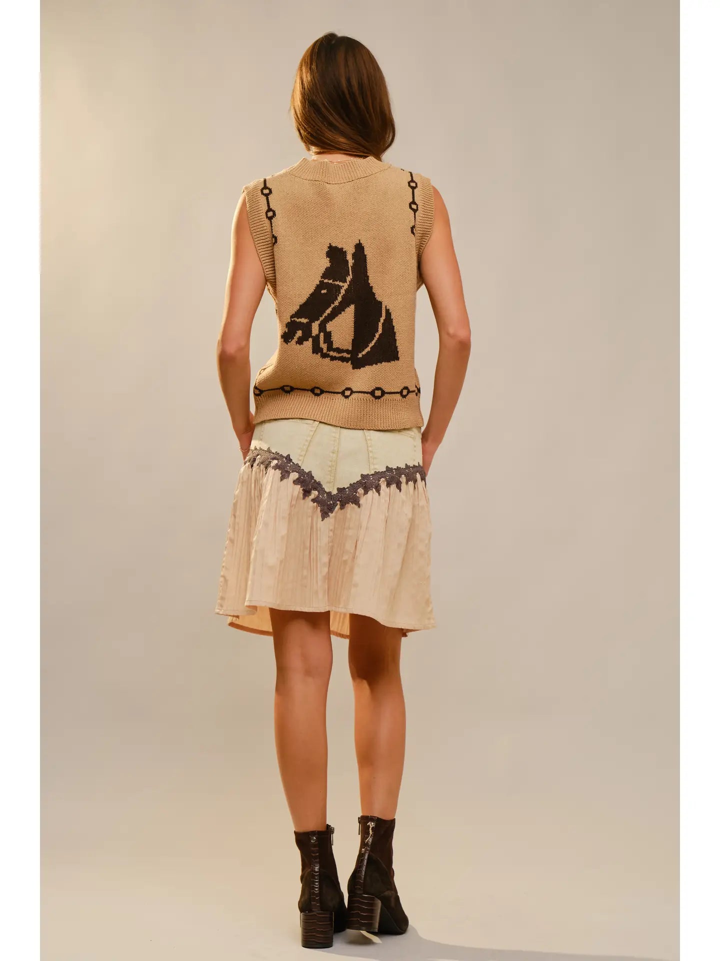 Back view of a woman wearing a beige sweater vest with a horse pattern and bit style chain 