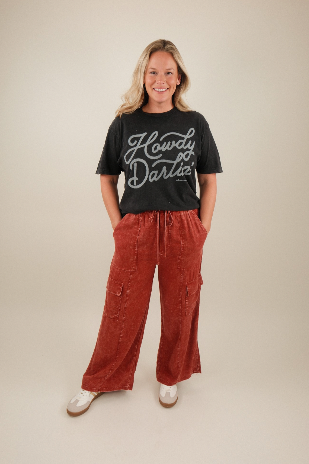 Woman wearing a black t-shirt with 'Howdy Darlin'' text and red cargo pants on a beige background