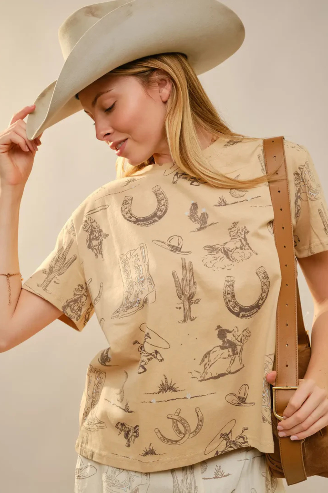 Woman wearing a beige shirt with western-themed designs and a cowboy hat, holding a brown leather bag.