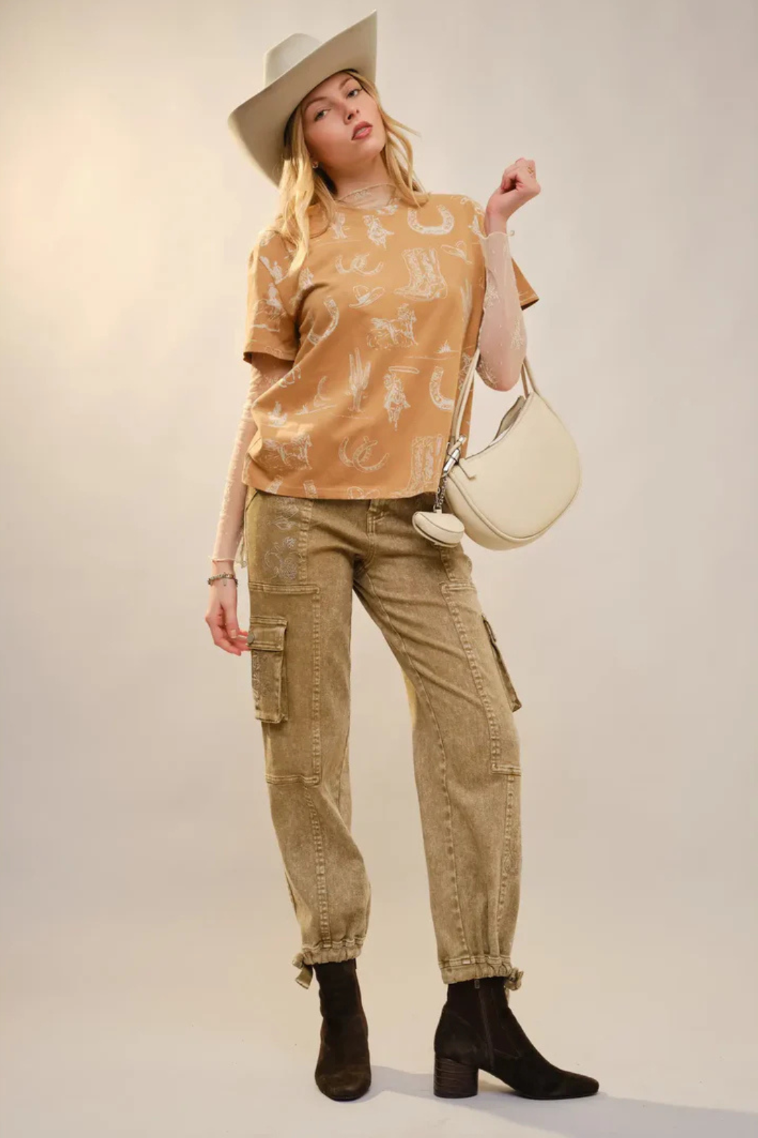 Woman wearing a camel western patterned top and beige cargo pants, holding a cream handbag against a plain background.