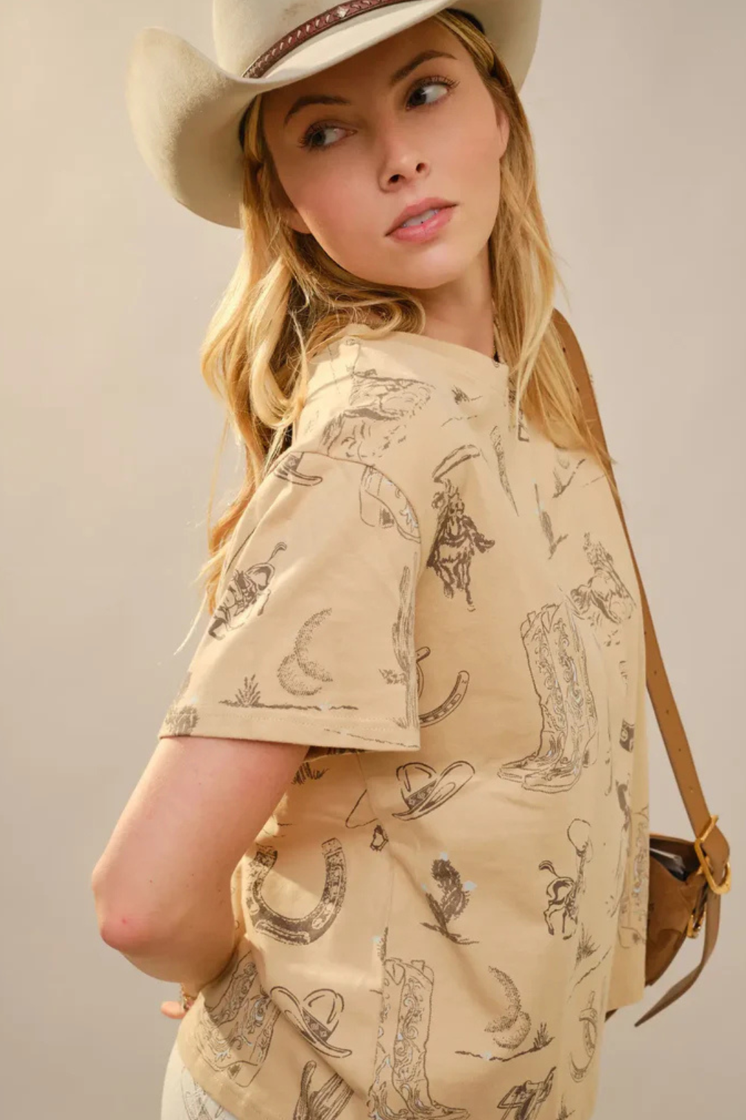 Woman wearing a beige cowboy hat and matching shirt with pattern, holding a brown handbag.