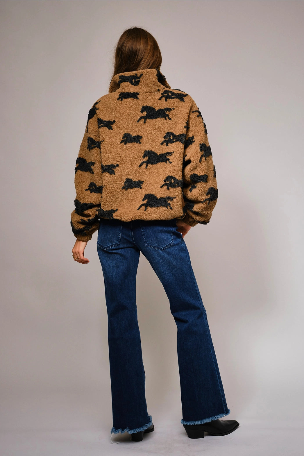 Person wearing a brown jacket with black horse pattern and blue jeans on a plain background