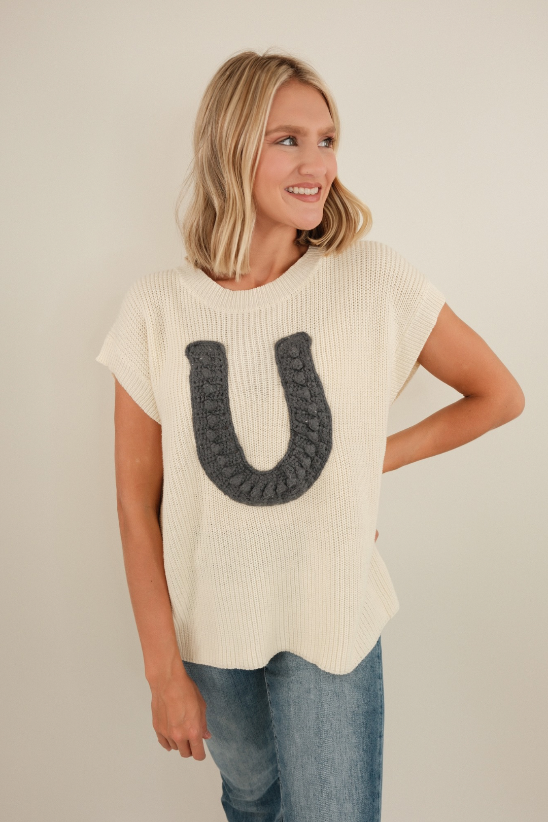 Chunky knit Wrangler’s Charm sweater in cream with ribbed sleeveless style and bold horseshoe motif to infuse cowgirl charm into any outfit.