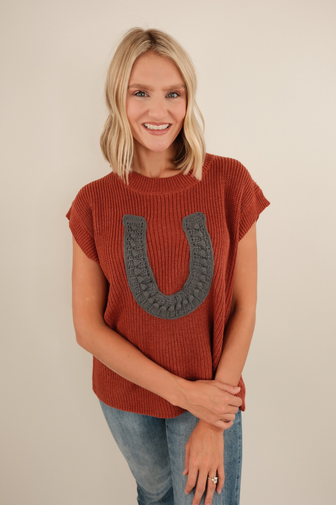 Chunky knit Wrangler’s Charm sweater in brick with ribbed sleeveless style and bold horseshoe motif to infuse cowgirl charm into any outfit