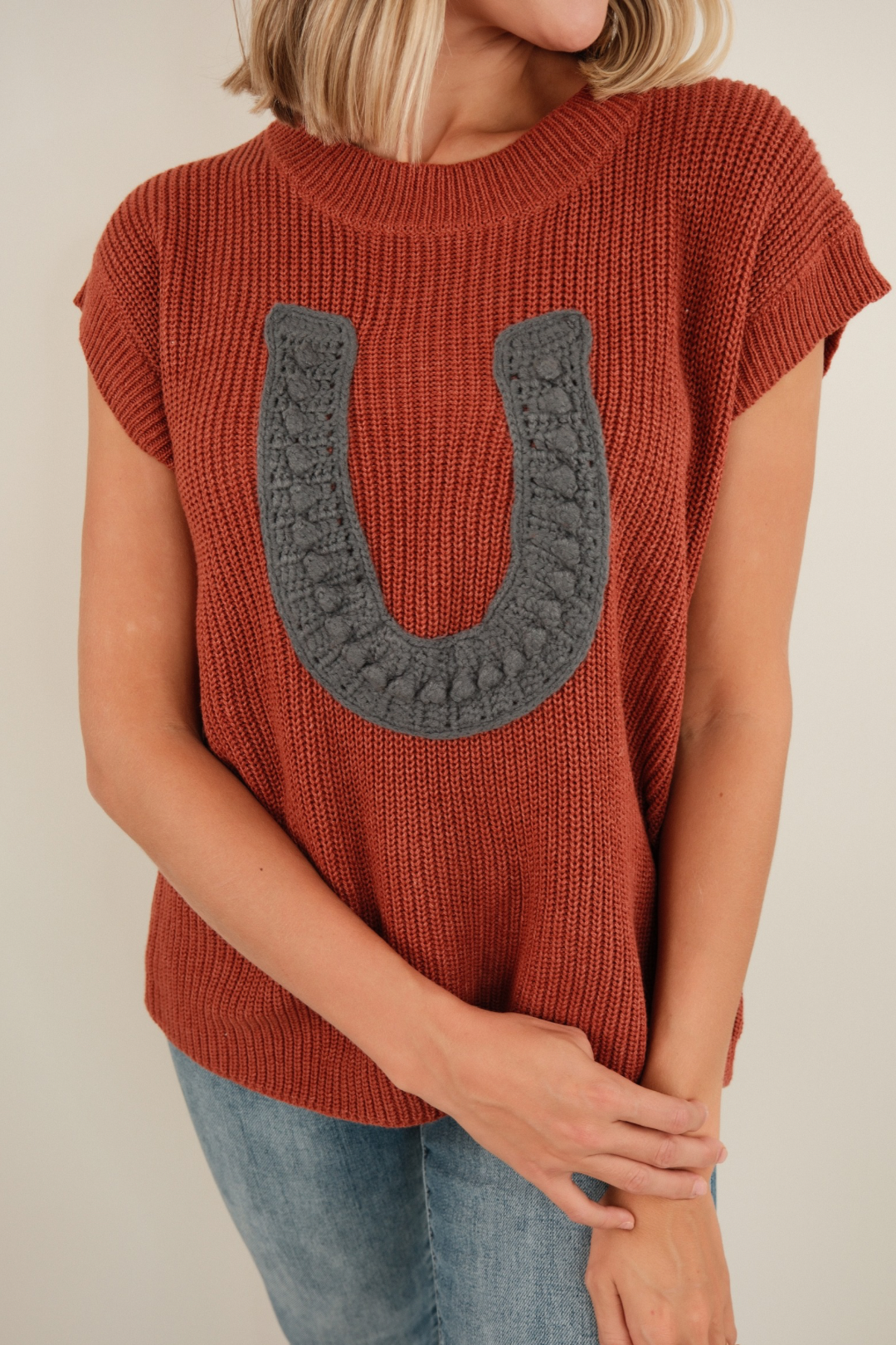 Front view of Wrangler’s Charm Sweater in brick, chunky ribbed sleeveless knit with bold horseshoe motif and western cowgirl style.